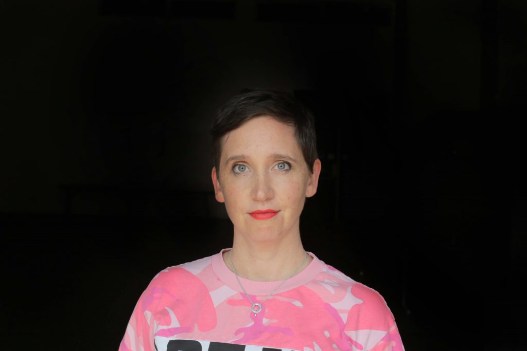 Dr Jessical Olivieri faces the camera against a black background wearing a pink shirt and red lipstick. She has short brown hair.