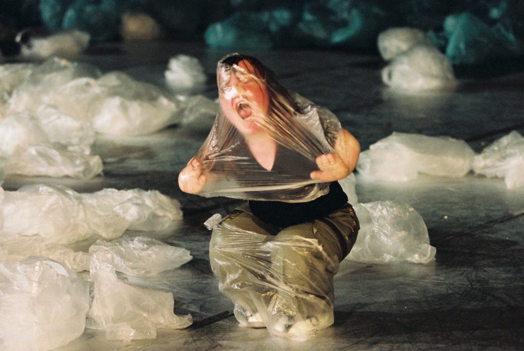 a woman screams, trapped head to toe in a clear plastic bag. she is crouching on a stage, surrounded by white plastic bags